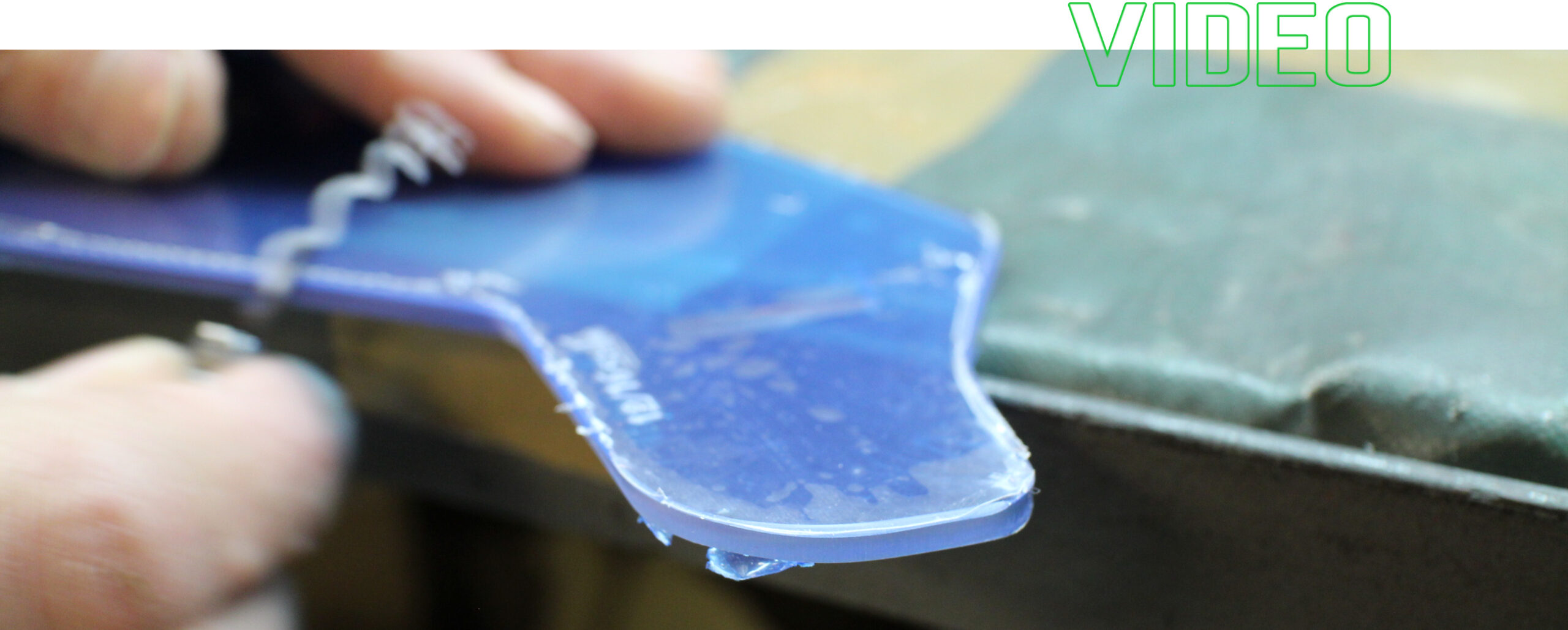 foto dettagliata delle mani di un operaio che leviga il bordo di una visiera blu in produzione. In alto a destra la scritta "VIDEO" contornata di verde