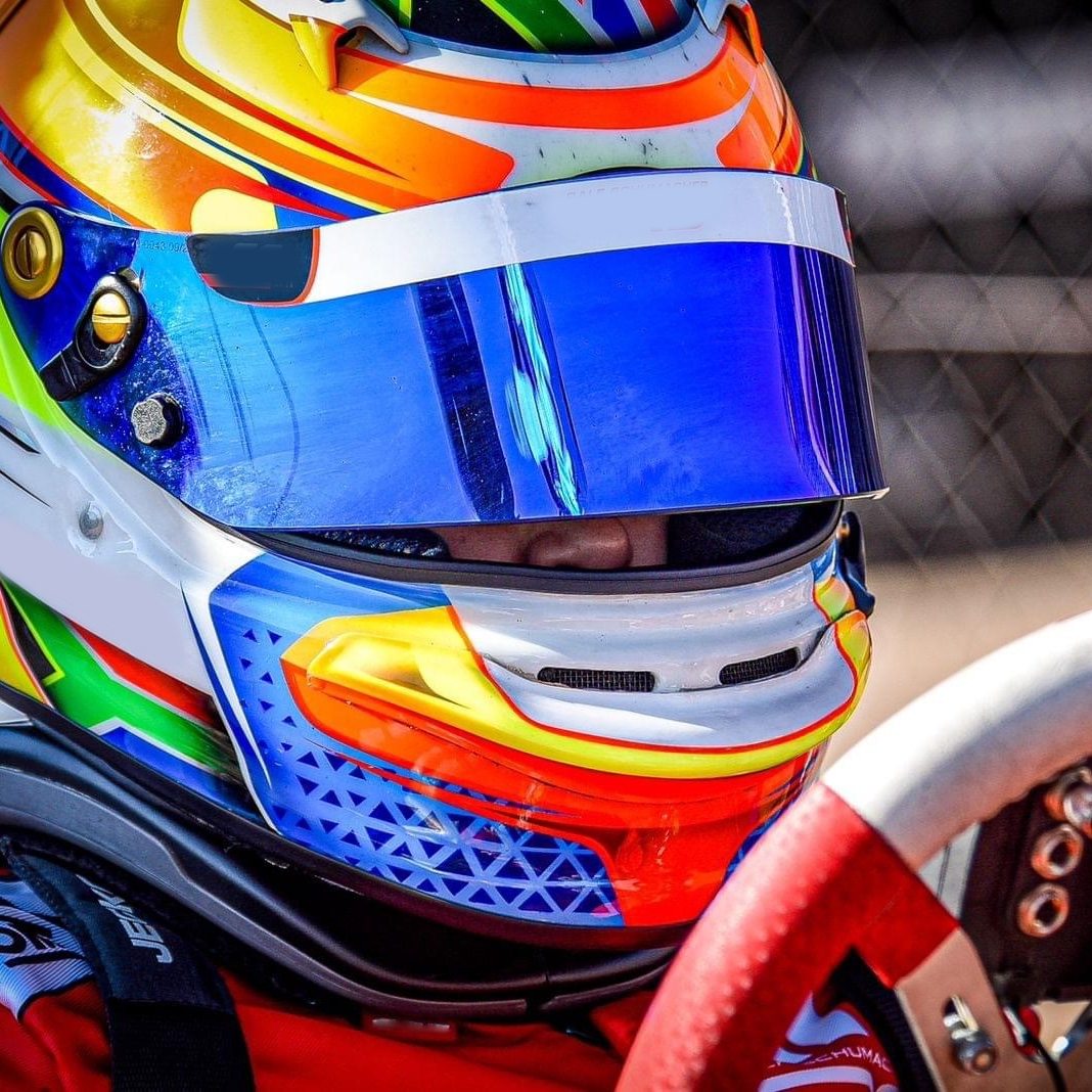 primo piano ravvicinato di un pilota di kart. vediamo molto bene il suo casco bianco con dettagli sgargianti arancioni, gialli e verdi. Ha una visiera blu con riflessi viola.