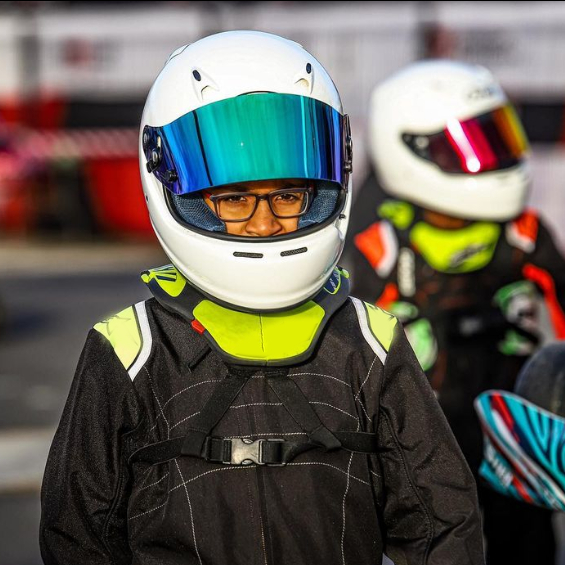 mezzo busto di un giovane pilota di kart, indossa una tuta nera e il suo casco è banco con la visiera blu con riflessi azzurri. la visiera è alzata e riusciamo a vedere gli occhi del giovane pilota. Lo sfondo è sfocato ma riusciamo a vedere la pista e un altro pilota vestito uguale e con lo stesso casco