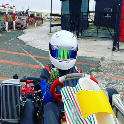 pilota di kart in pista parcheggiato. Indossa un casco bianco con visiera blu con riflessi verdi. Ha una tuta nera e blu e il suo kart è bianco con strisce verdi e rosse.