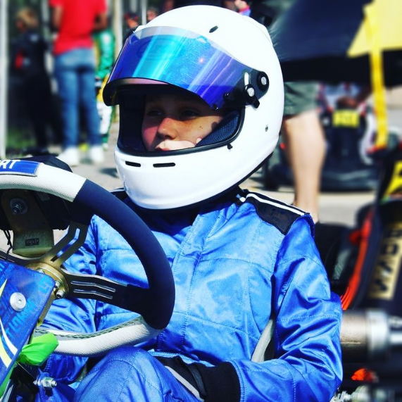 primo piano di un pilota di kart a riposo nel suo veicolo. ha una visiera blu con casco bianco e tuta azzurra. Dietro di lui, sfondo sfocato ma si intravedono altri piloti fermi come lui